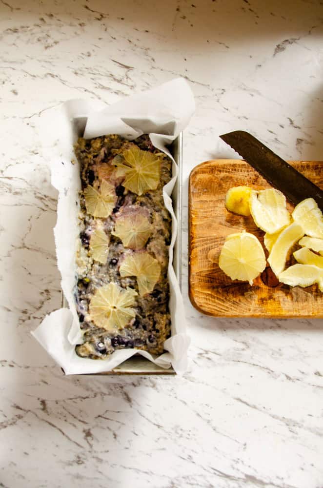 A loaf pan with parchment paper and blueberry lemon batter and sliced lemons on top with a cutting board beside it with fresh cut lemons.