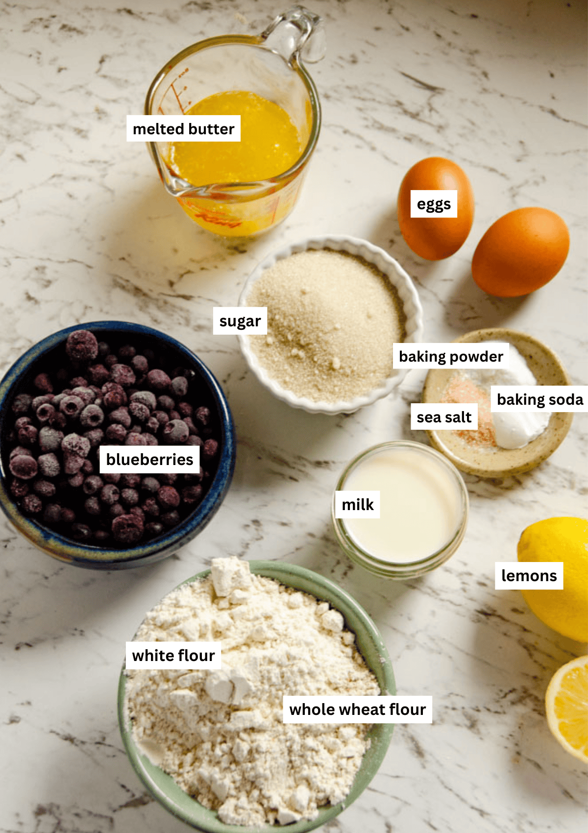 Labelled ingredients for blueberry lemon load including melted butter, eggs, sugar, milk, lemons, baking soda, baking powder, sea salt, blueberries, whole wheat and white flour.