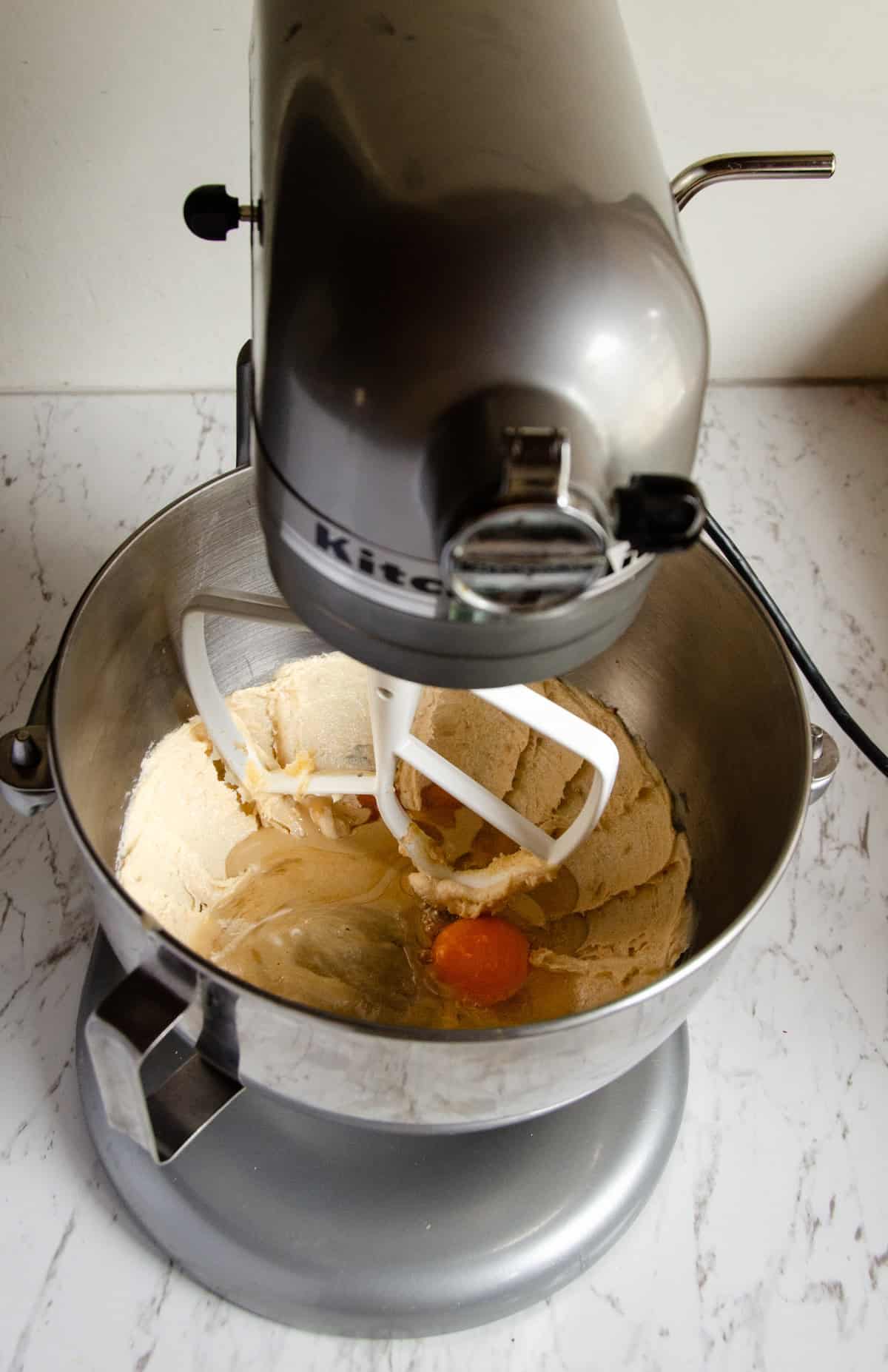 A mixing bowl with a paddle attachment with batter and an egg just cracked into the bowl.