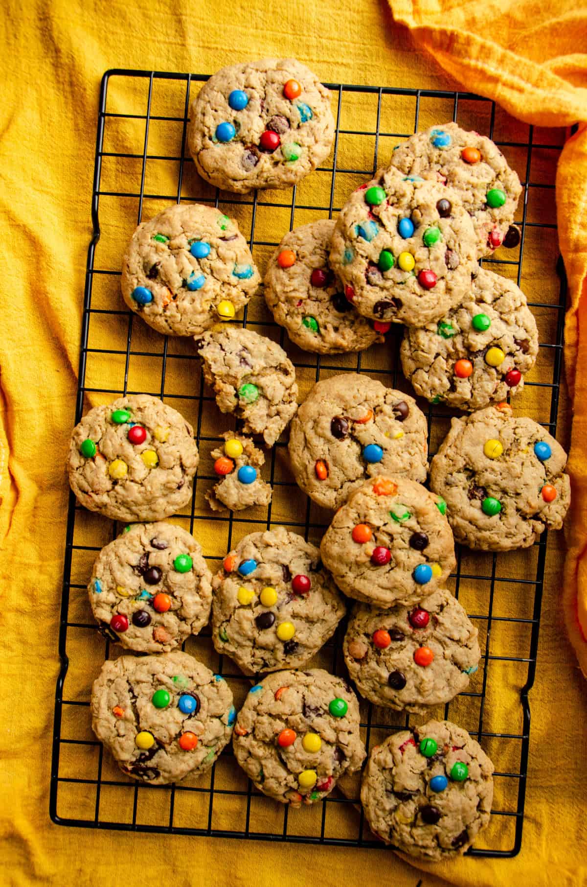 A cooling rack covered with light brown cookies that have specks of oats, chocolate chips and colorful chocolate candies.