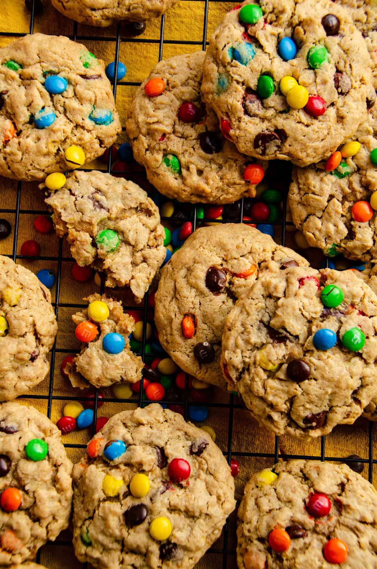 A cooling rack covered with overlapping light brown cookies that have specks of oats, chocolate chips and colorful chocolate candies.