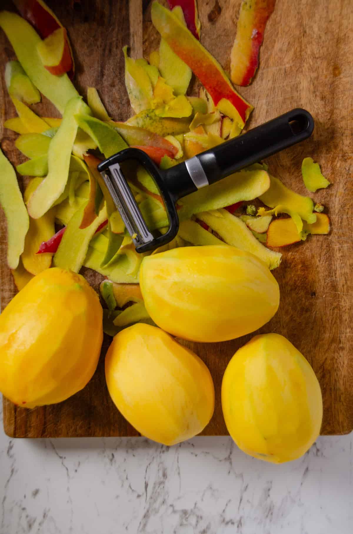 Mangoes on a cutting board that have been peeled with a peeler and peels beside it.