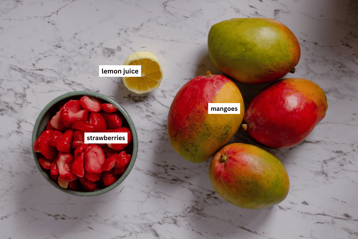 Chopped strawberries in a bowl, half a lemon, and 4 large mangoes.