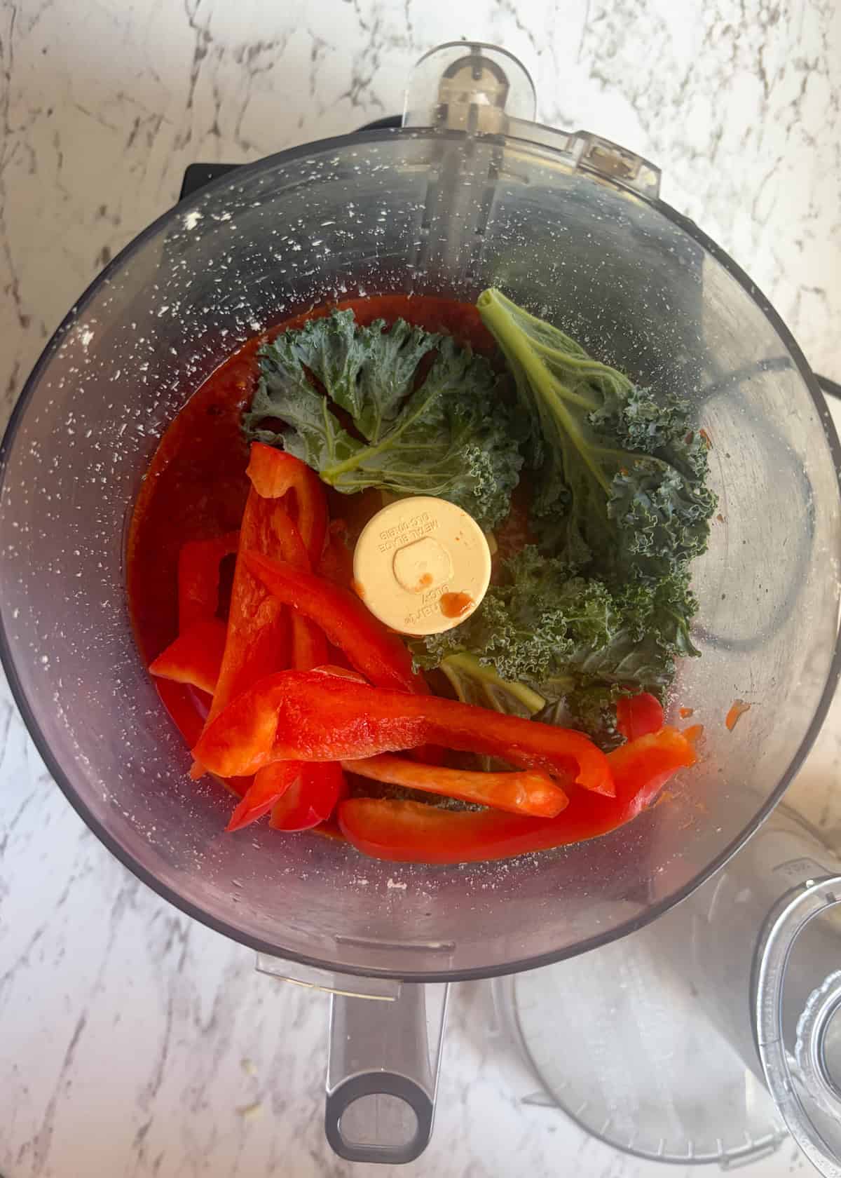 A food processor with red sauce, red peppers, and kale in it.