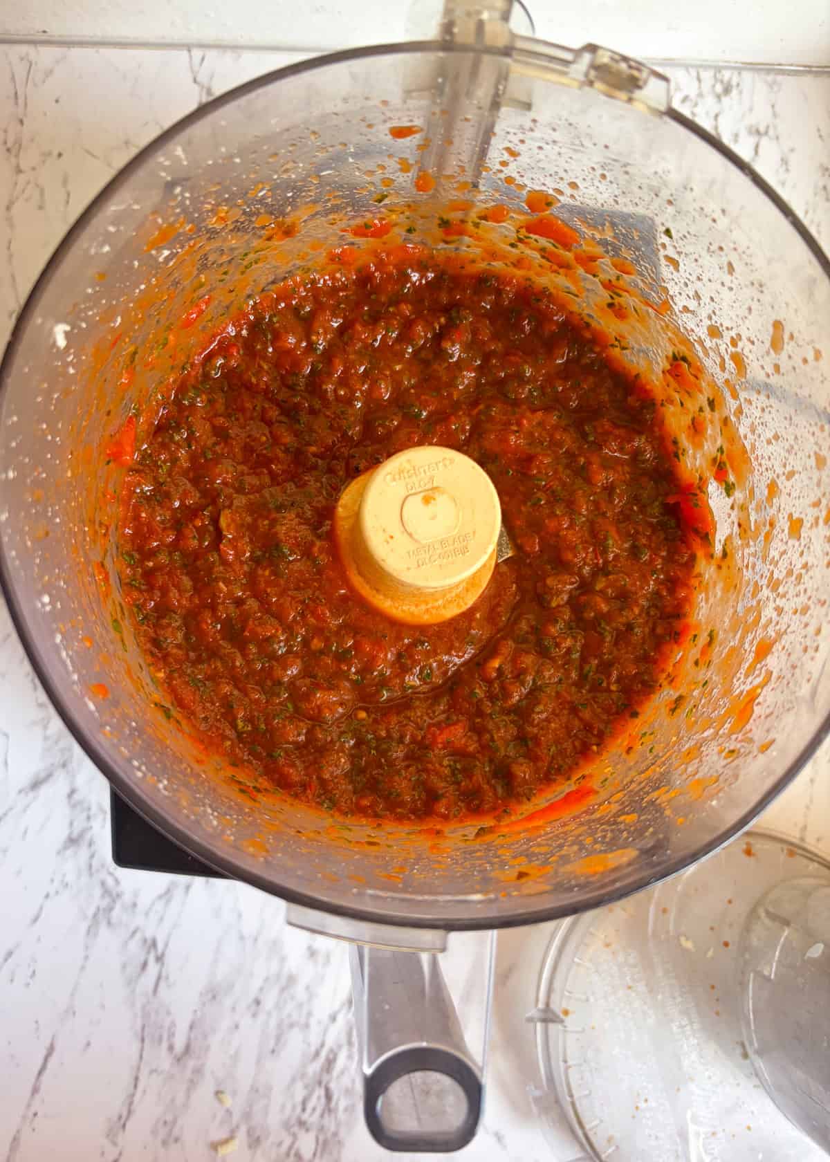 A food processor with red sauce in it with green flecks from kale.