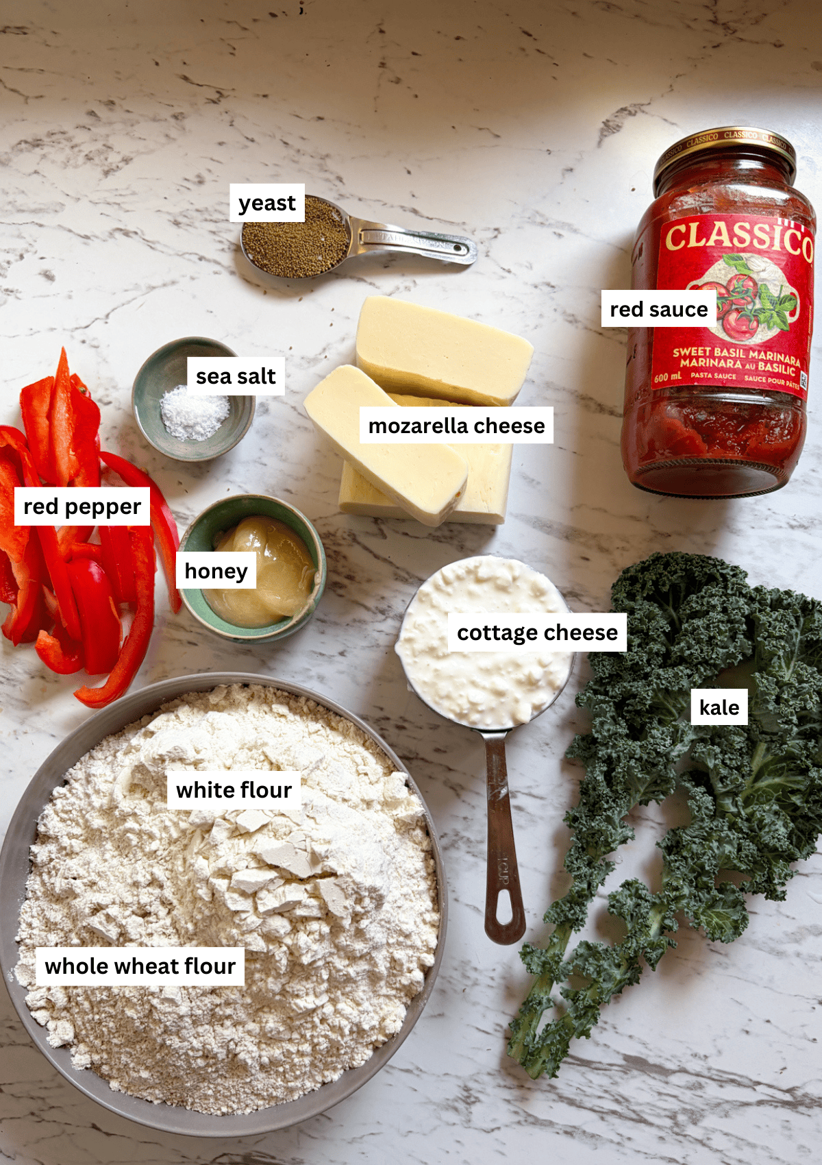 Ingredients for pizza rolls out on the counter including red pepper, kale, flour, cottage cheese, honey, sea salt, yeast, mozarella cheese, and red sauce.