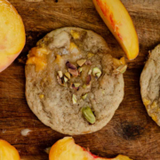 A cookie on a wooden tray with chopped pistachios on top and sliced peaches beside it.