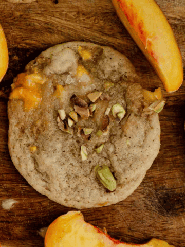 A cookie on a wooden tray with chopped pistachios on top and sliced peaches beside it.