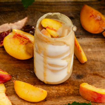 A peach smoothie in a glass cup with yogurt streaks on the inside and peaches all around it.