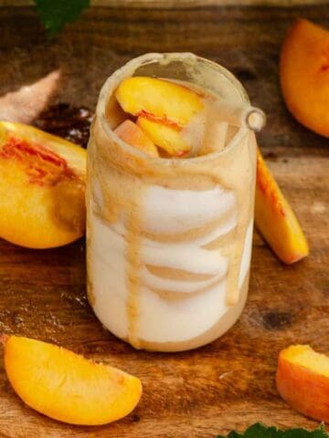 A peach smoothie in a glass cup with yogurt streaks on the inside and peaches all around it.