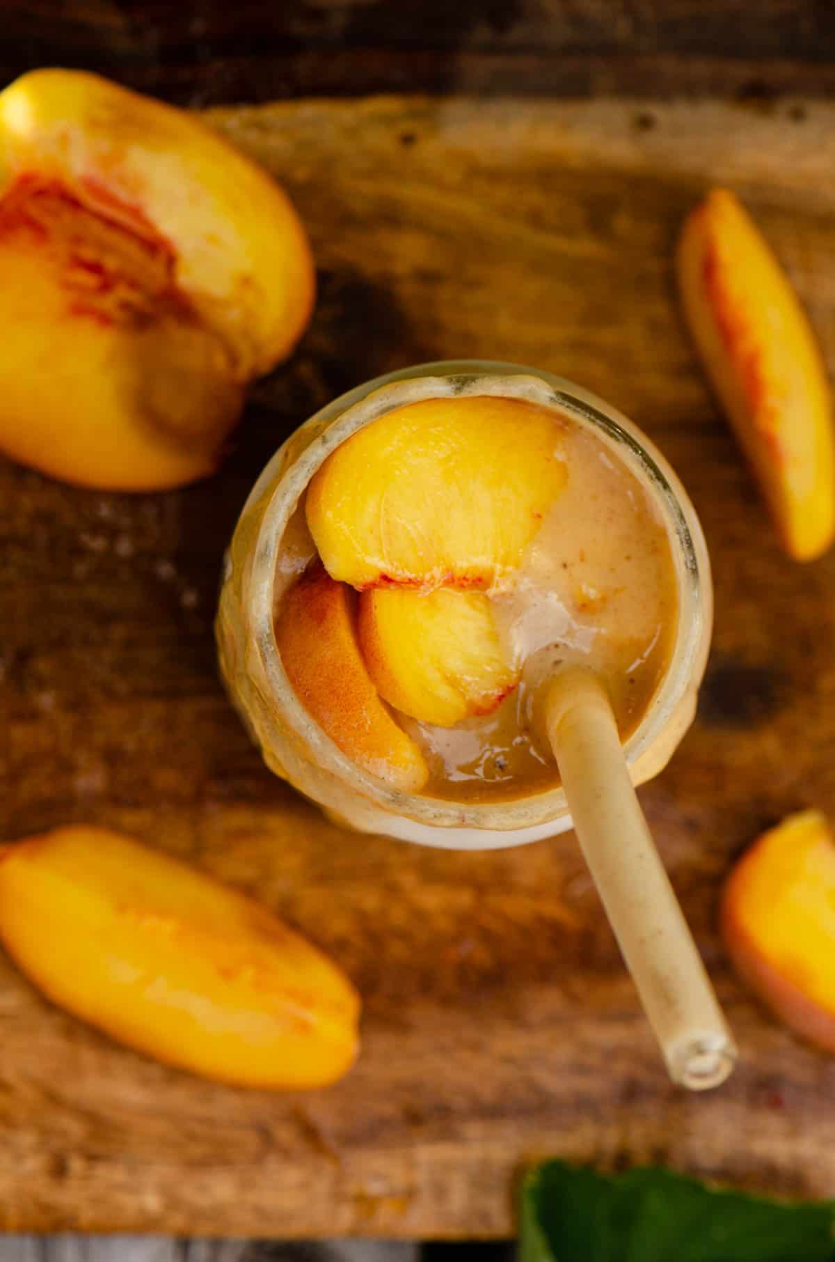 A light peach-colored smoothie in a glass cup with two sliced peaches on top and a glass straw in it, with a sip taken out of the cup as the cup is not full .