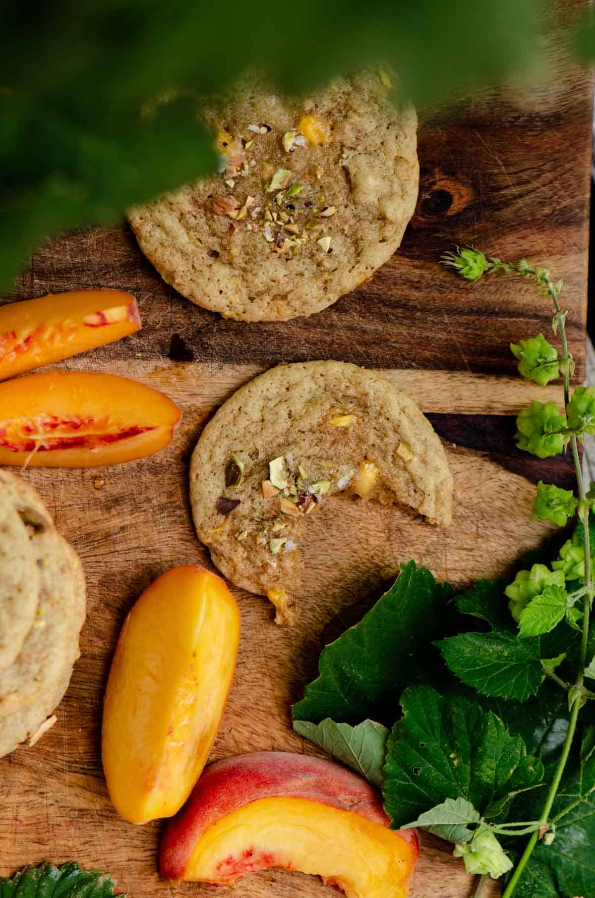 A cookie on a wooden sheet with a bite out of it with chop pistachios on top and sliced peaches beside it.