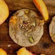 A cookie on a wooden tray with chopped pistachios on top and sliced peaches beside it.