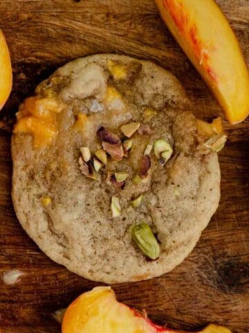 A cookie on a wooden tray with chopped pistachios on top and sliced peaches beside it.