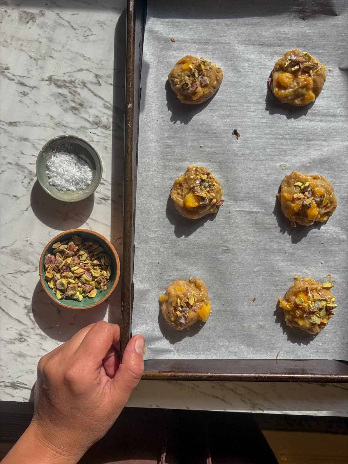 Cookie dough balls on a parchment covered cookie sheet being sprinkled with chunky salt and chopped pistachios.