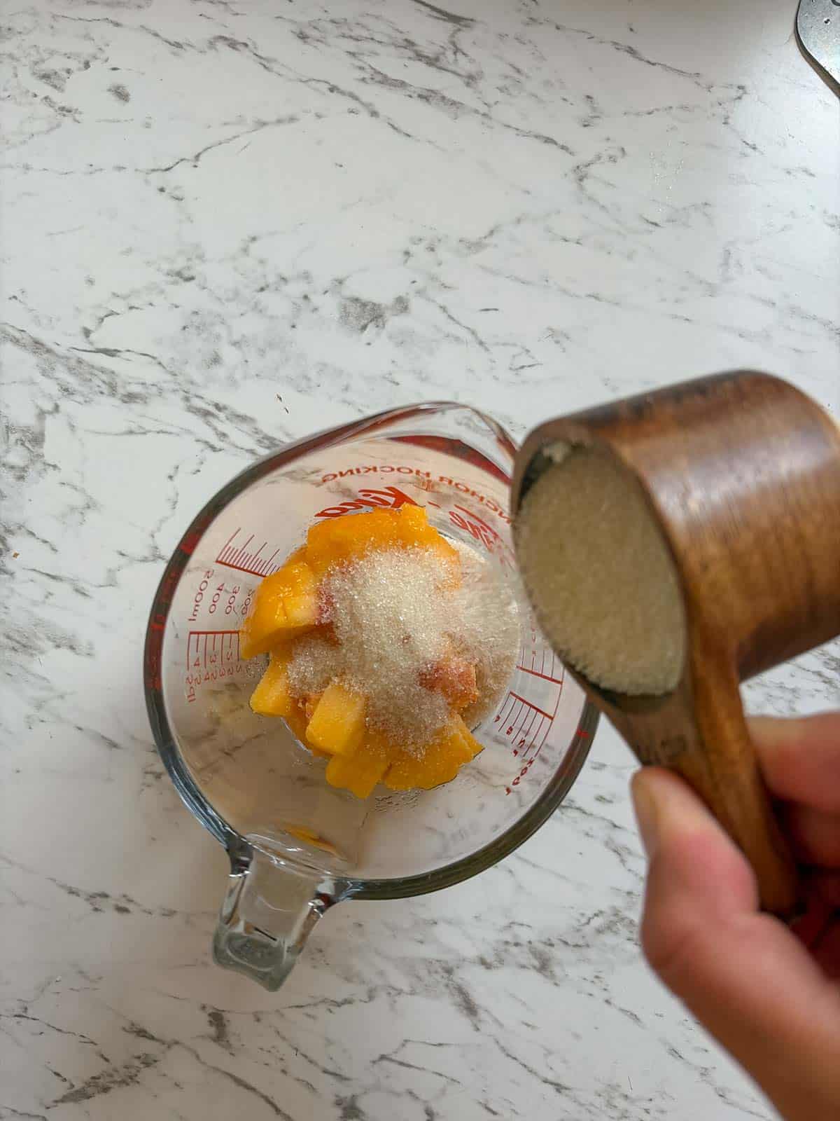 A glass measuring cup with chopped peached in it, with a hand pouring white sugar onto the peaches.